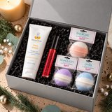 Open silver gift box with Marigoldbub Face Polish, Glossberry lip oil and three packaged Roxy Grace bath bombs, surrounded by greenery and warm candlelight.